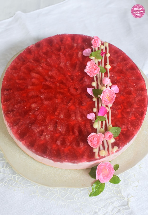Himbeer-Cheesecake auf rohweißer Steingutplatte dekoriert mit Streifen weißer Schokolade, grünen Zitronenmelisseblättern und kleinen rosa Röschen