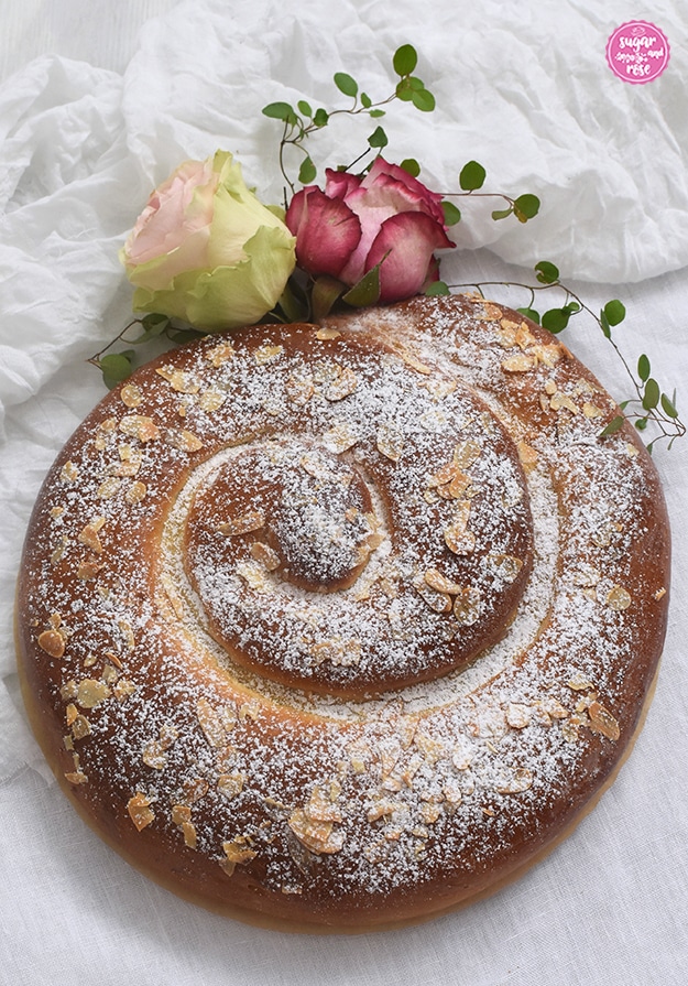 Große Apfelstrudel-Zimtschnecke auf weißem Leinentischtuch, dekoriert mit zwei Rosen, eine dezent hellrosa mit grünen Außenblättern, die andere rosa mit kräftig rotem Rand