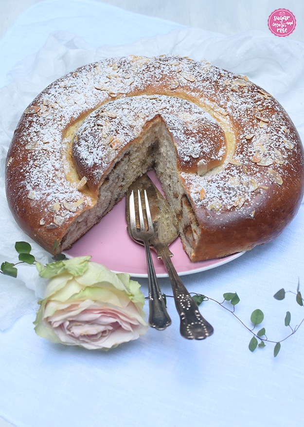 Apfel-Zimtschnecke auf rosa Keramikteller, ein großes Stück ist herausgeschnitten, man sieht im Anschnitt den braunen Zimt. Am Teller liegt eine silberne Tortenschaufel und eine Dessertgabel