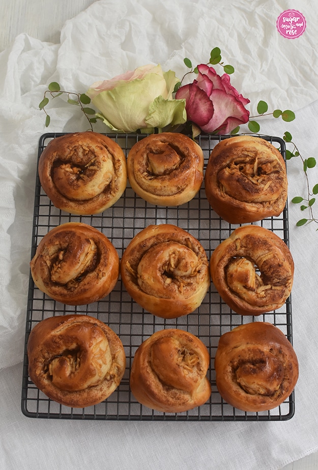 Neun kleine Apfel-Zimtschnecken auf einem dunkelgrauen Gitter zum Abkühlen, dekoriert mit zwei rosa Rosen und grünen Blättern