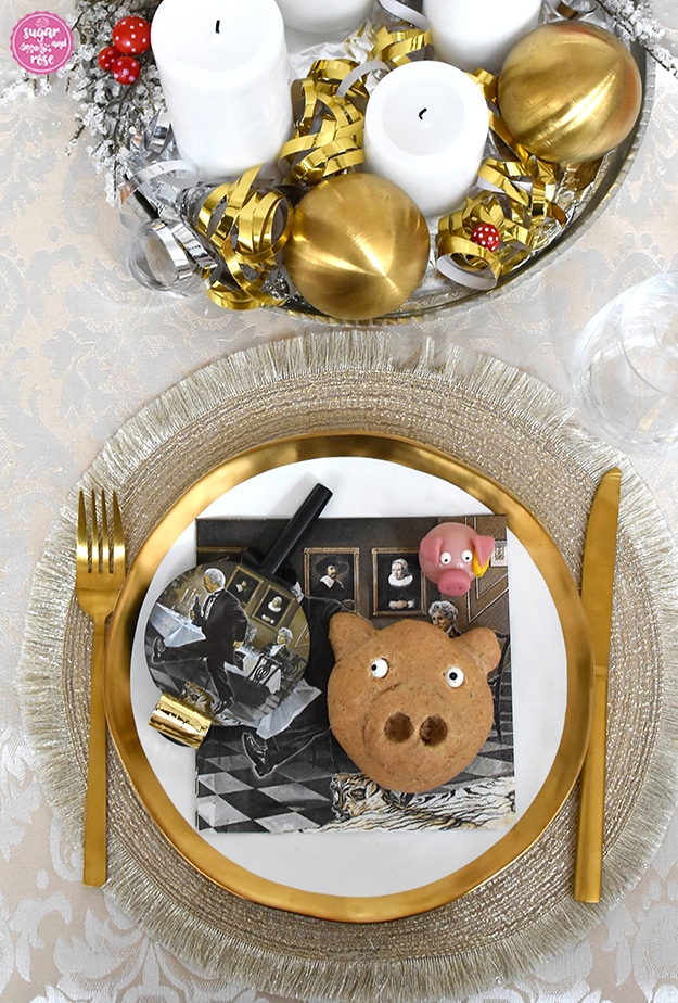 Glücksschweinderl-Brötchen auf Teller mit Goldrand, daneben Goldbesteck und ein goldfarbenes Platzset, im Hintergrund ein silberner Teller mit vier weißen Kerzen, zwei Goldkugeln und silber und goldene Luftschlangen