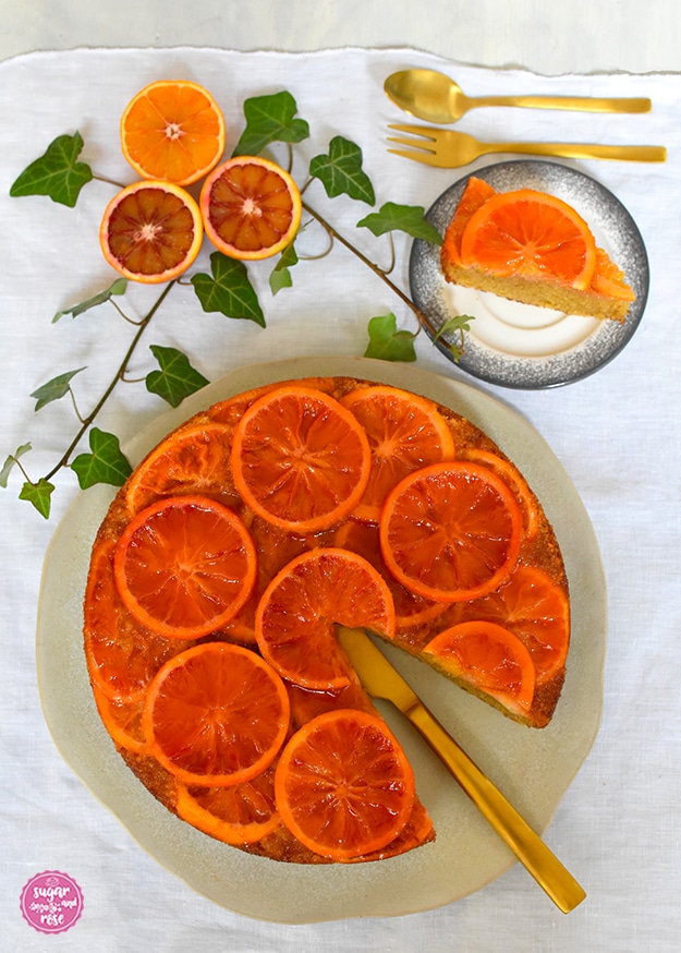 Angeschnittener Blutorangenkuchen auf Keramikplatte, dahinter Blutorangenhälften mit unterschiedlichen Färbungen, ein Stück Kuchen auf einem grau-weißen Gmunder-Keramik-Teller, dazu goldenes Dessertbesteck (Löffel und Gabel), daneben Efeuranken