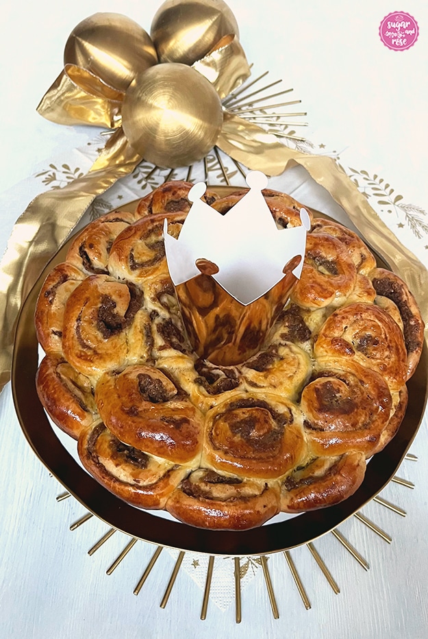 Dreikönigskuchen mit Kletzen auf Goldrandteller, in der Kuchenmitte eine goldene Papierkrone, dahinter drei goldene Dekokugeln und eine Goldmasche