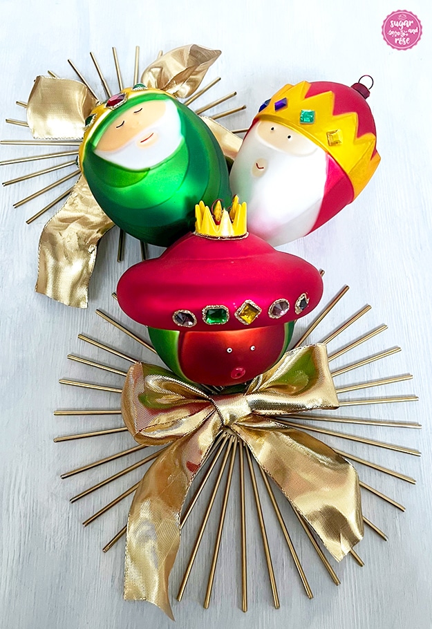 Drei bunte Alessi-Glaskugel-Weihnachtsbaumanhänger mit den Köpfen der drei Könige in rot, grün und gold-rot, liegend auf zwei sonnenförmigen goldenen Untersetzern