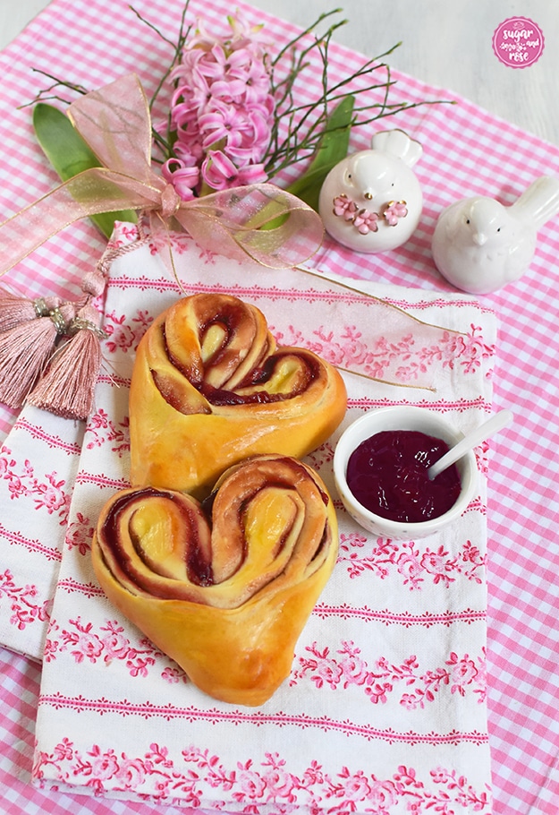 Zwei Himbeer-Hefeteigherzen auf rosa kariertem und mit rosa Rosen bedruckten Geschirrtüchern, dahinter eine rosa Hyazintenblüte und zwei kleine weiße Porzellantäubchen