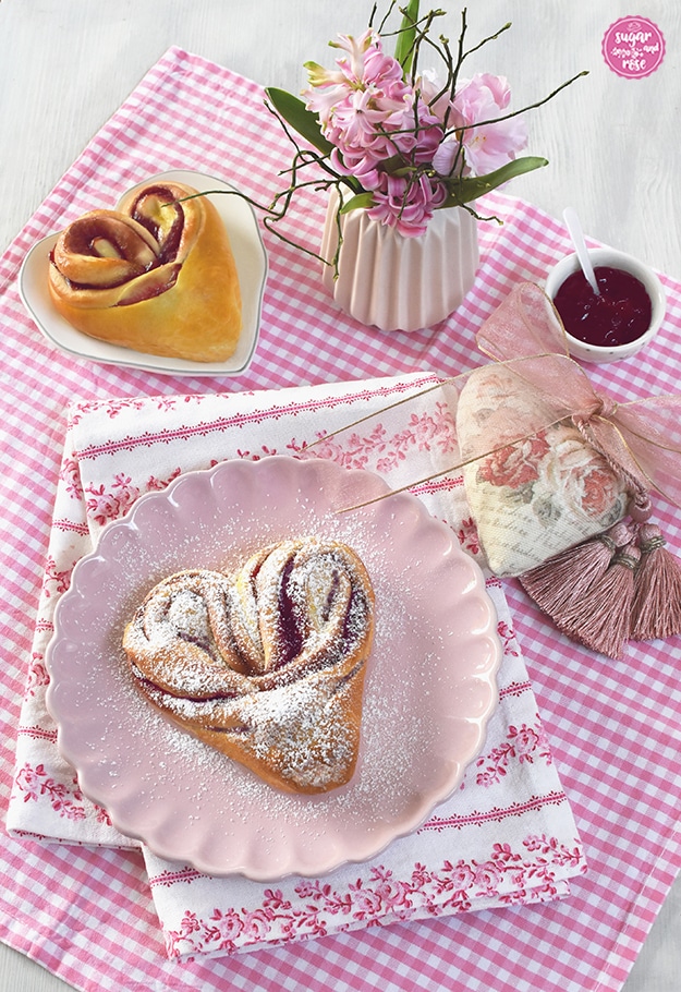 Ein gezuckertes Himbeer-Hefeteigherz auf rosa Keramikteller im Vordergrund, daneben ein Vintage-Stoffherz mit Rosenblütendekor und rosa durchsichtiger Masche, dahinter ein Himbeer-Hefeherz auf herzförmigen Teller und eine kleine Vase mit Hyazintenblüten
