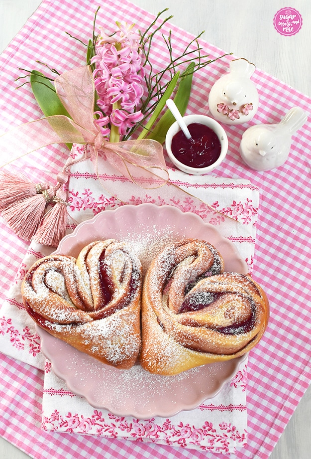 Zwei Himbeer-Hefeteigherzen mit Puderzucker auf einem rosa Porzellanteller, darunter ein rosa kariertes und ein mit rosa Rosen bedrucktes Geschirrtuch, dahinter eine rosa Hyazintenblüte und zwei kleine weiße Porzellantäubchen sowie eine kleine Schale mit Himbeergelee