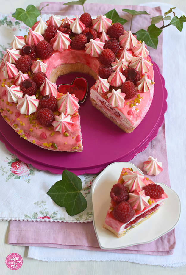 Himbeer-Pistazien-Saturntorte angeschnitten auf rosa Tortenteller, im Vordergrund ein herzförmiger Desserteller mit einem Stück Torte