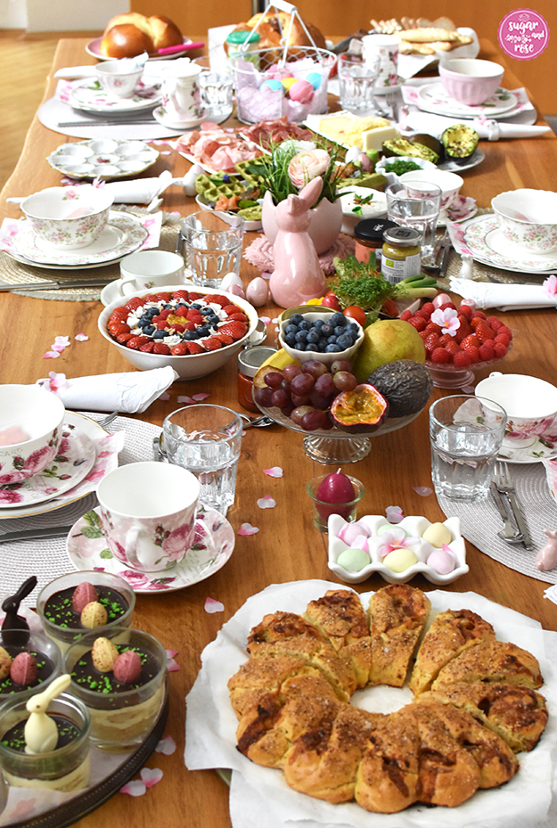 Osterbrunch-Tisch gedeckt mit rosa Rosen-Porzellan, Osterschinken, bunten Ostereiern, einer Schale Müsli, diverse Früchte und Beeren, Spinatwaffeln mit Lachs, im Vordergrund ein rundes Pizzabrot und Bananenschnitten in Dessertgläsern