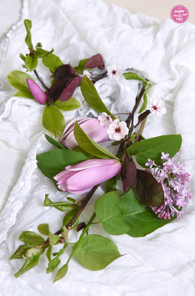 Zwei Zweige mit fliederrosa Tulpenmagnolienblüten, ein Zweig mit weißen Blutpflaumenblüten mit zarten weinroten Staubgefäßen und weinroten Blättern sowie eine helllila Fliederblütenrispe auf weißem Tuch mit zarter weißer Spitze