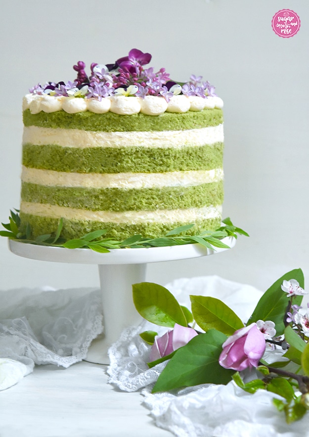 Matcha-Eierlikör-Torte auf weißer Etagere in Frontalansicht, davor rosa Magnolienblüten und eine weiße Spitzendecke