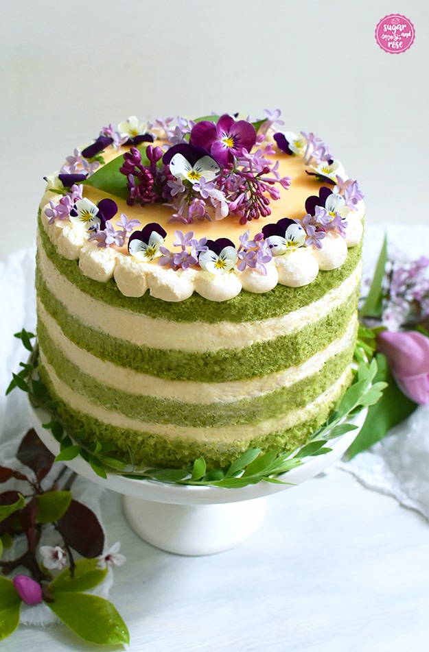Matcha-Eierlikör-Torte mit vier grünen Biskuitböden und drei weiß-gelben Eierlikörcremeschichten, dekoriert mit Cremetupfen und violett-weißen Violen