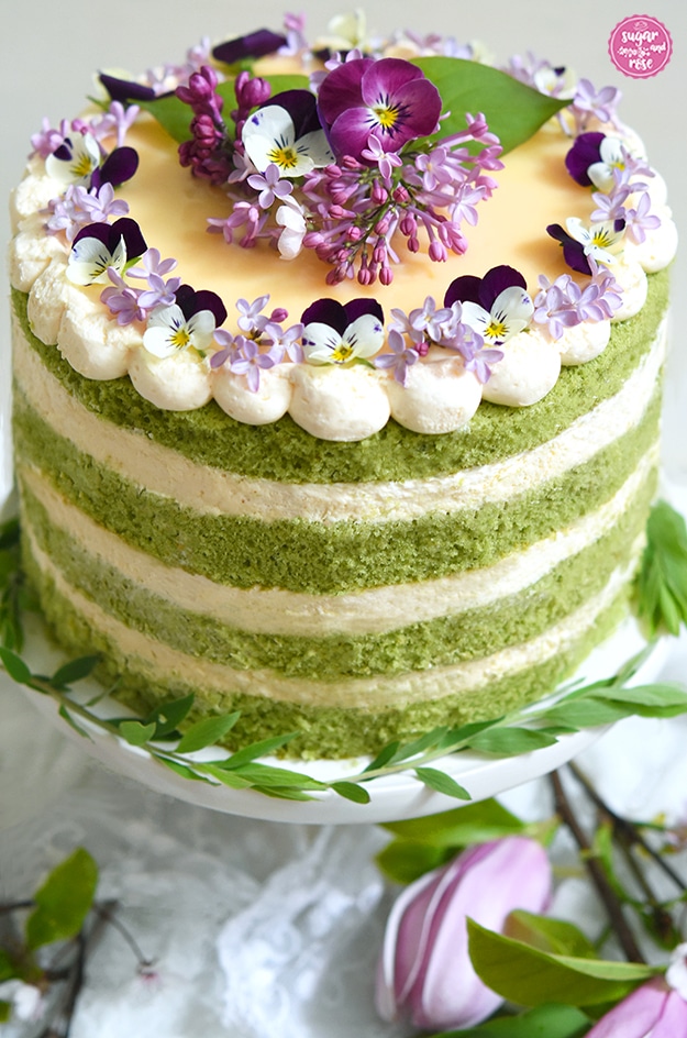 Matcha-Eierlikör-Torte von oben mit sichtbarem gelbem Eierlikörspiegel zwischen den hellen Cremetupfen; in der Tortenmitte eine helllila Fliederrispe, grüne Blätter und zwei Hornveilchenblüten in unterschiedlichen Fuchsia-, Dunkelviolett-Tönen mit weißen und gelben Mitten