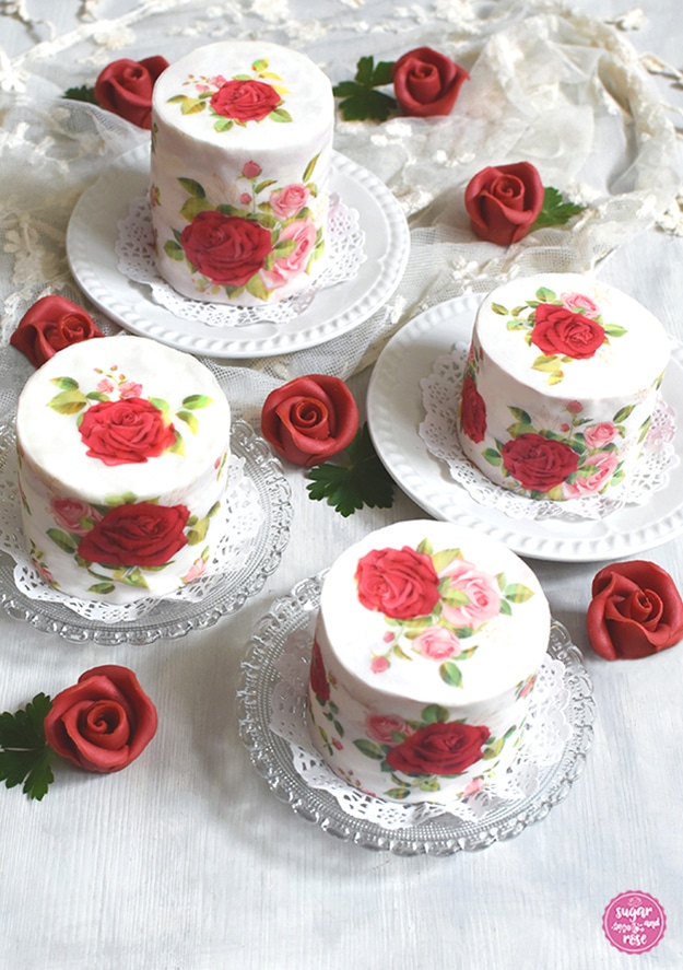 Vier kleine runde Rosentörtchen mit unterschiedlicher Höhe, jedes mit Rosenmotiven in rot und rosa bedruckt, dazwischen mehrere dunkelrote Marzipanrosenblüten und grüne Blütenblätter