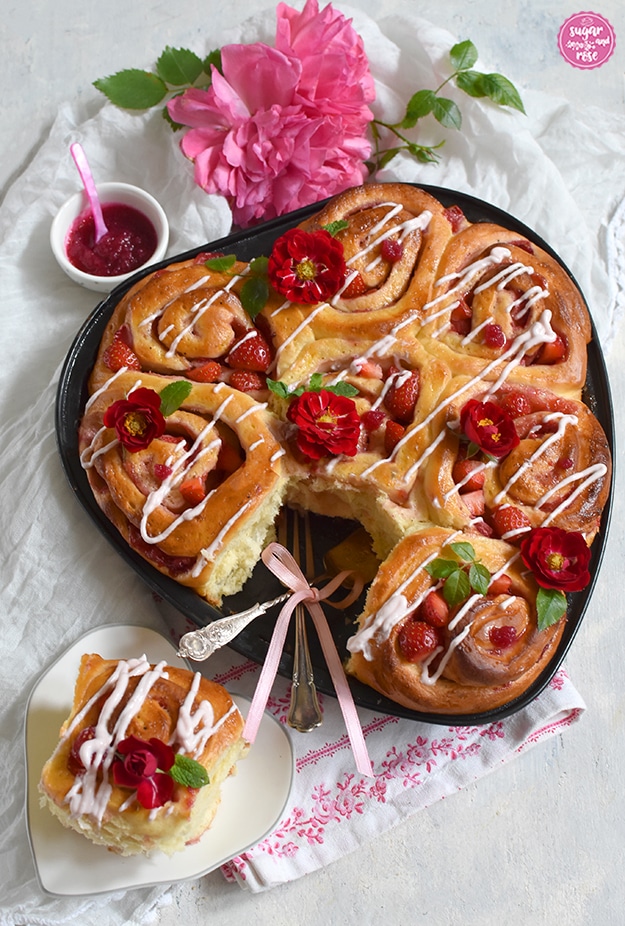 Erdbeerherz gebacken und mit Frischkäsestreifen und kleinen roten Röschen der Rose Rote The Fairy dekoriert, im Vordergrund eine Hefeschnecke auf einem weißen herzförmigen Keramikteller