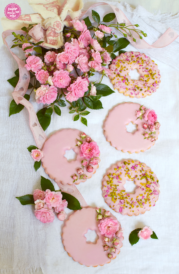 Fünf Rosenkekse, davon drei mit kleinen Röschen bestreut, die anderen zwei mit grünen Pistazien und pinken Rosenblütenschnipseln, dekoriert mit einem Strauß rosa Rosen