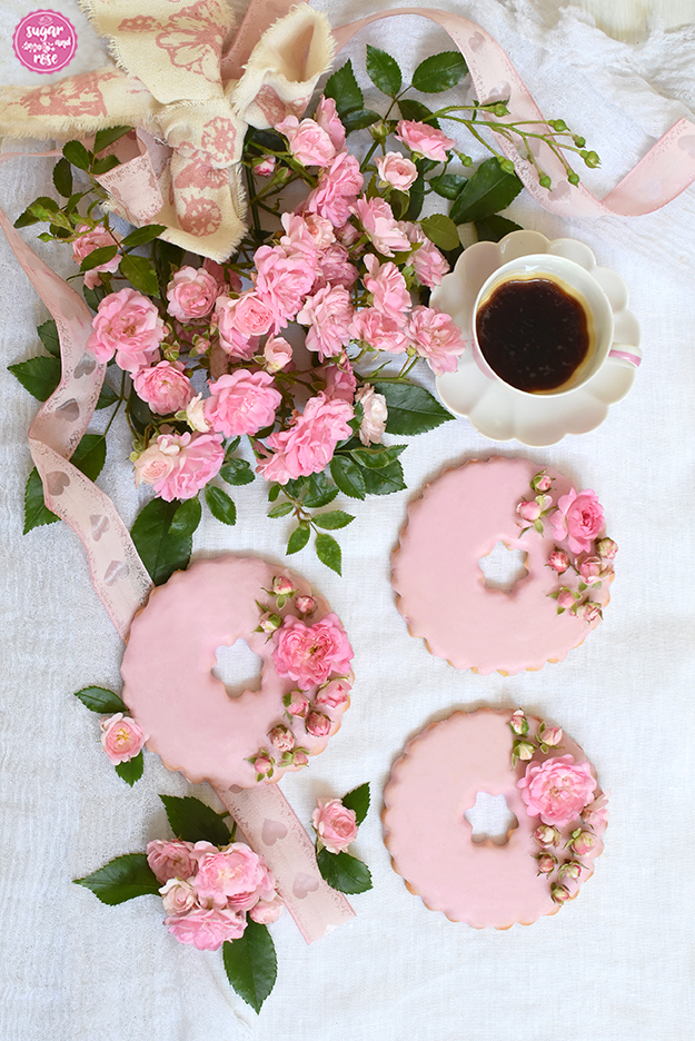 Drei große rosa Rosenkekse mit kleinen rosa Rosenblüten auf weißem Leinentuch mit einer kleinen rosa Moccatasse von Augarten, daneben ein Strauß rosa Röslein mit rosa Schleife