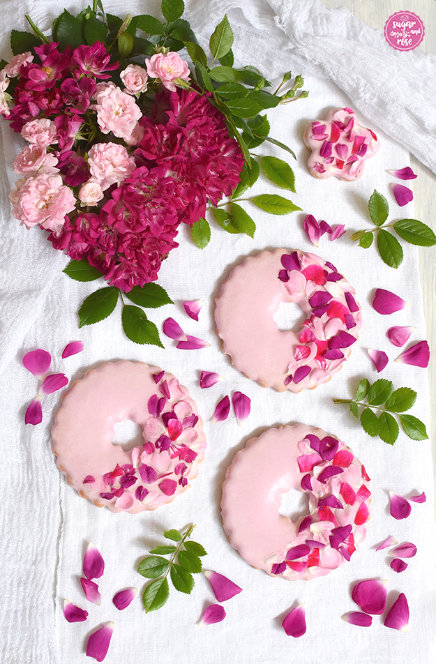 Drei rosa Kekse mit Loch, zur Hälfte dekoriert mit verschiedenfarbigen Rosenblüten in Fuchsia, Pink und Hellrosa