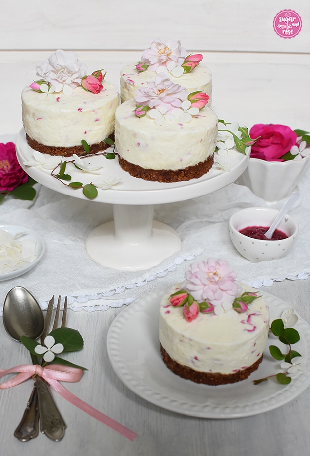 Ein kleiner Rosen-Kokos-Cheesecake auf einem weißen Vintage-Keramikteller im Vordergrund, drei auf einer kleinen weißen Etagere im Hintergrund, daneben silber Dessertgabel und Löffel, die Cheesecakes sind mit kleinen blassrosa Rosenblüten und dunkelrosa Knospen dekoriert