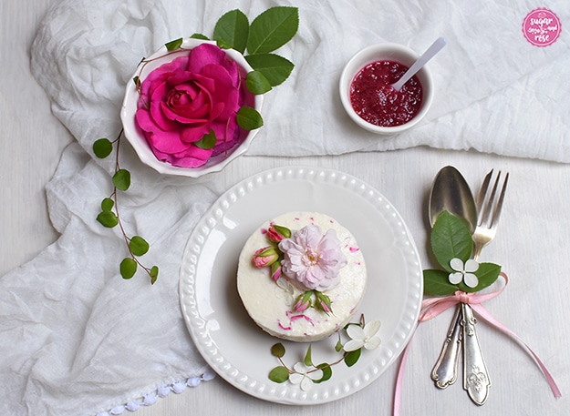 Frozen Rosen-Kokos-Cheesecake auf weißem Dessertteller, dekoriert mit einem blassrosa Röschen, rosa Rosenknospen und weißen kleinen Blüten; daneben altsilbernes Dessertbesteck, eine Schale mit einer pinken Rosenblüte und ein Schälchen mit rotem Rosenmus in Minilöffelchen