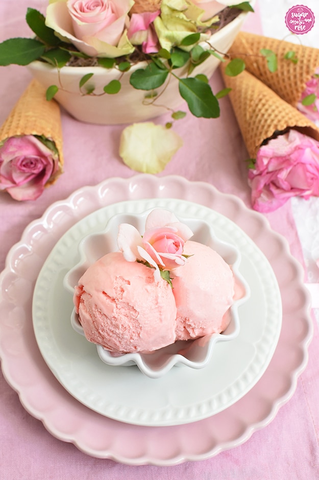 Zwei Kugeln Rosen-Eis in kleiner weißer Keramikschale auf weißem und rosa Teller, darunter eine rosa Leinenserviette, im Hintergrund eine ovale Schale mit rosa und weiß-grünen Rosenblüten, daneben Eiswaffeltüten mit rosa Rosen