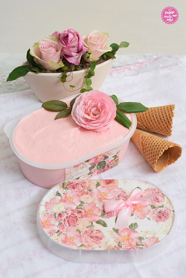 Große ovale Kunststoffbox gefüllt mit Roseneis und dekoriert mit einer Rosenblüte, die Box ist ebenso wie der davor liegende Deckel mit Rosenblütenpapier dekoriert, daneben Waffeltüten, dahinter Schale mit drei rosa Rosen