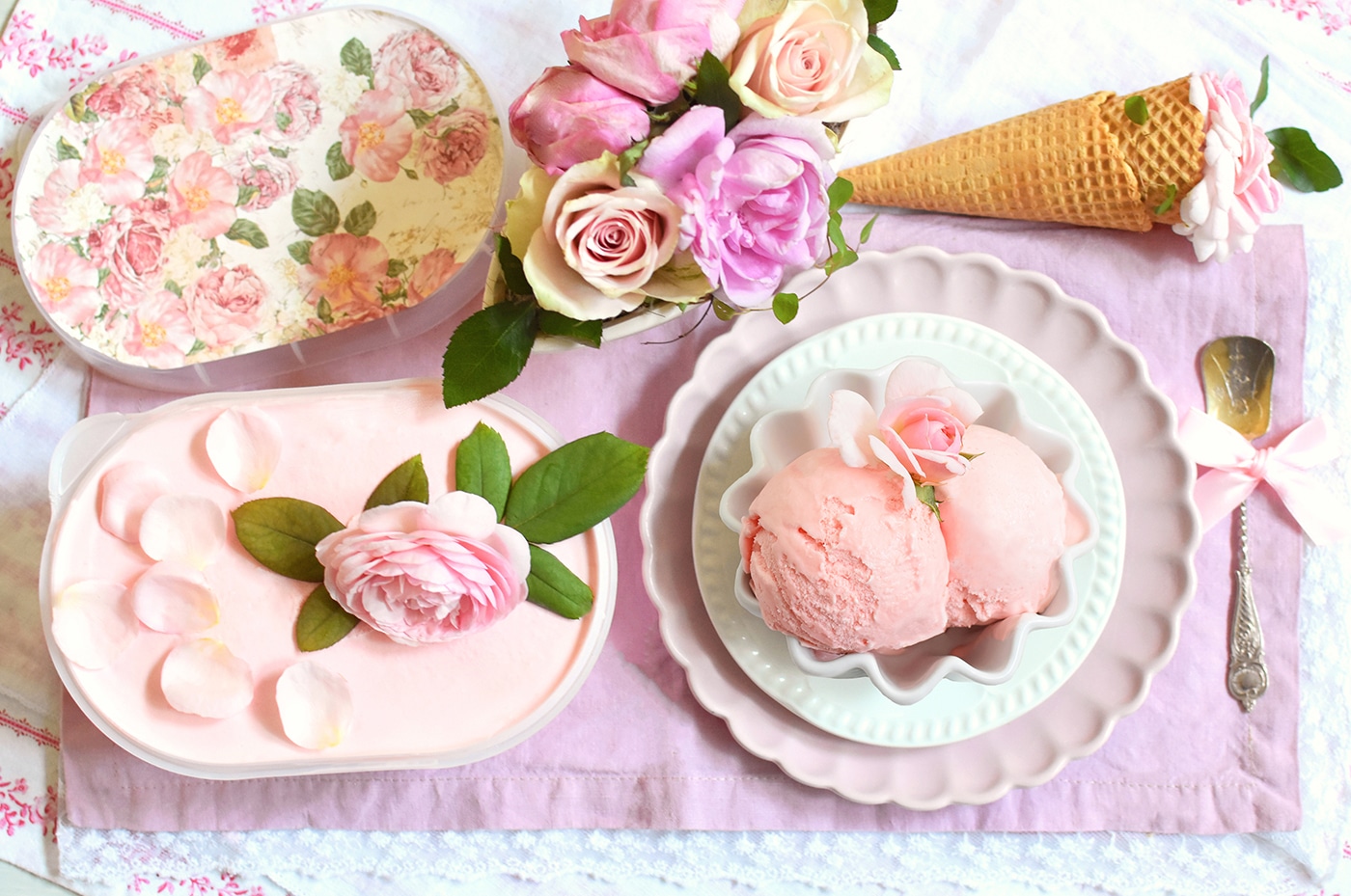 Hellrosa-farbiges Roseneis in ovaler Box, dekoriert mit Rosenblüten, daneben eine Schale mit zwei Kugeln Roseneis, im Hintergrund zwei Waffel-Cones und eine Schale mit mehreren rosa Rosenblüten