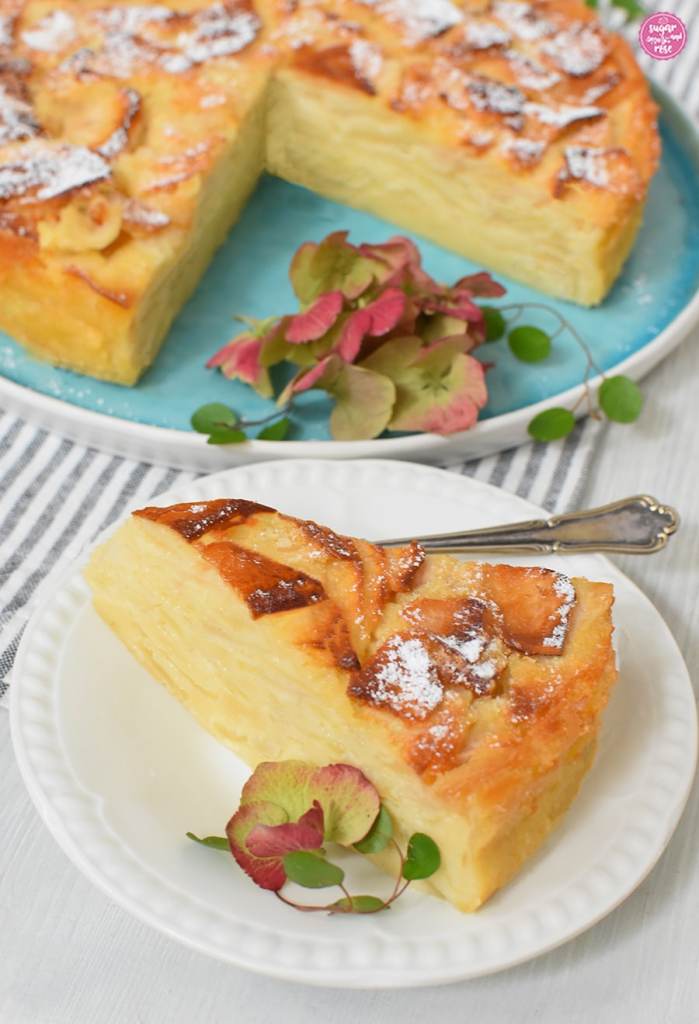 Ein Stück und dahinter der angeschnittene Apfelkuchen, dekoriert mit rosa Hortensienblütw und grünen Blättern