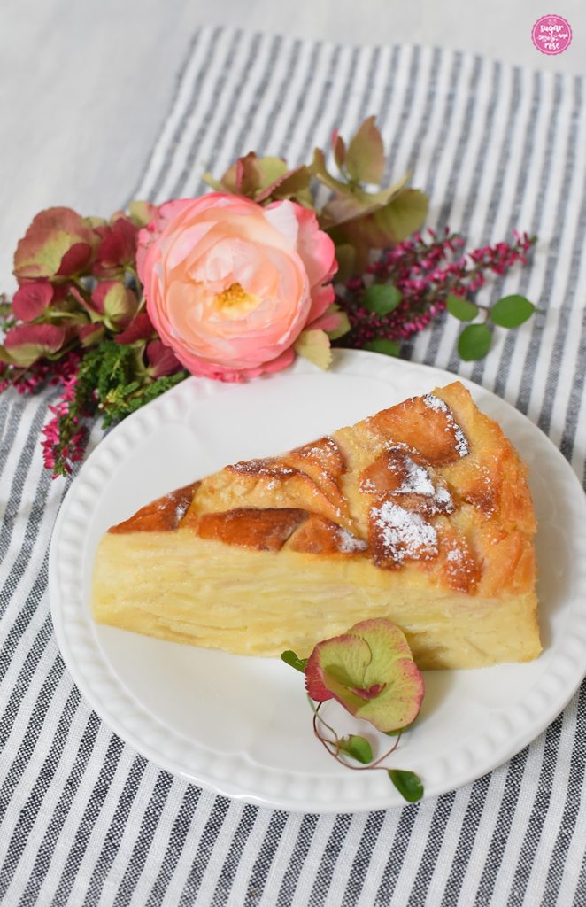 Apfelkuchen all'italiana Kuchenstück auf weißem Vintage-Teller mit einer einzelnen grün-rosa Hortensienblüte, dahinter eine Blüte der cremeweiß-rosa Nostalgirose, Hortensien und rosa Heidekraut