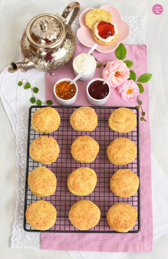 Zwölf Scones auf einem schwarzen Gitterrost dahinter eine silberne Teekanne und drei Schälchen gefüllt mit Clotted Cream, Erdbeermarmelade und Orangenmarmelade