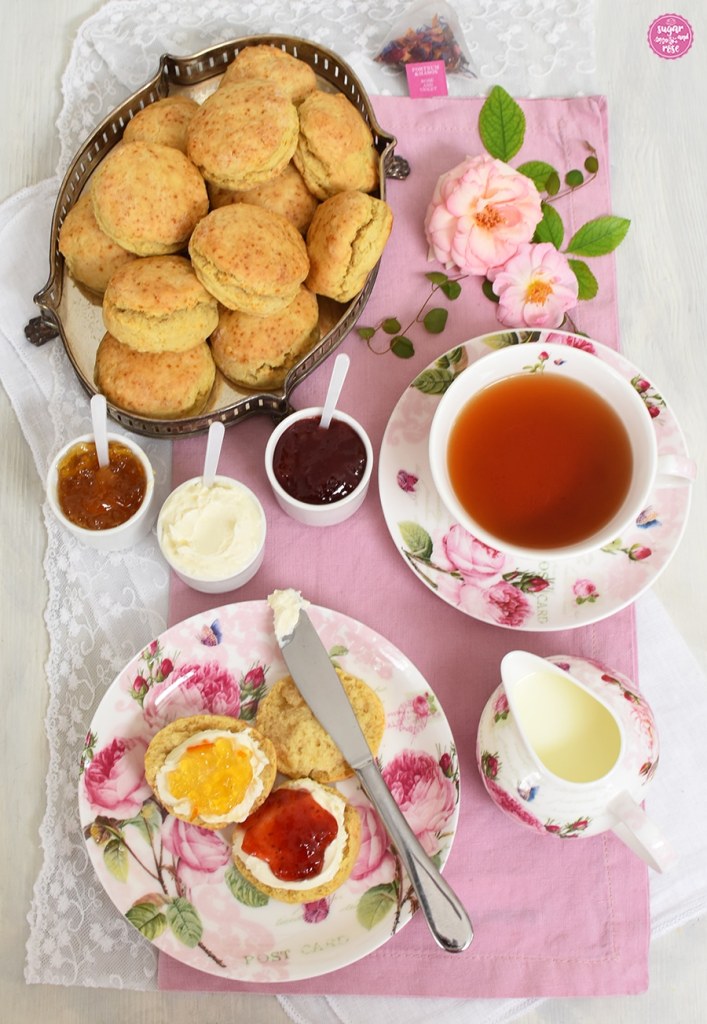 Eine Tasse Tee, eine Schale mit Milch sowie ein Teller mit aufgeschnittenen und bestrichenen Scones, ein Messer, dahinter eine Silberschale mit zahlreichen Scones sowie Marmelade und Clotted Cream Schälchen