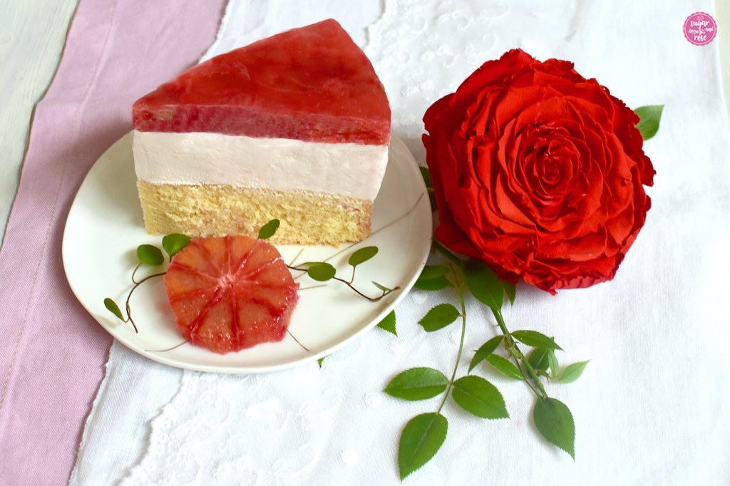 Tortenstück auf weißem Dessertteller mit Blutorangenscheibe und roter Rosenblüte