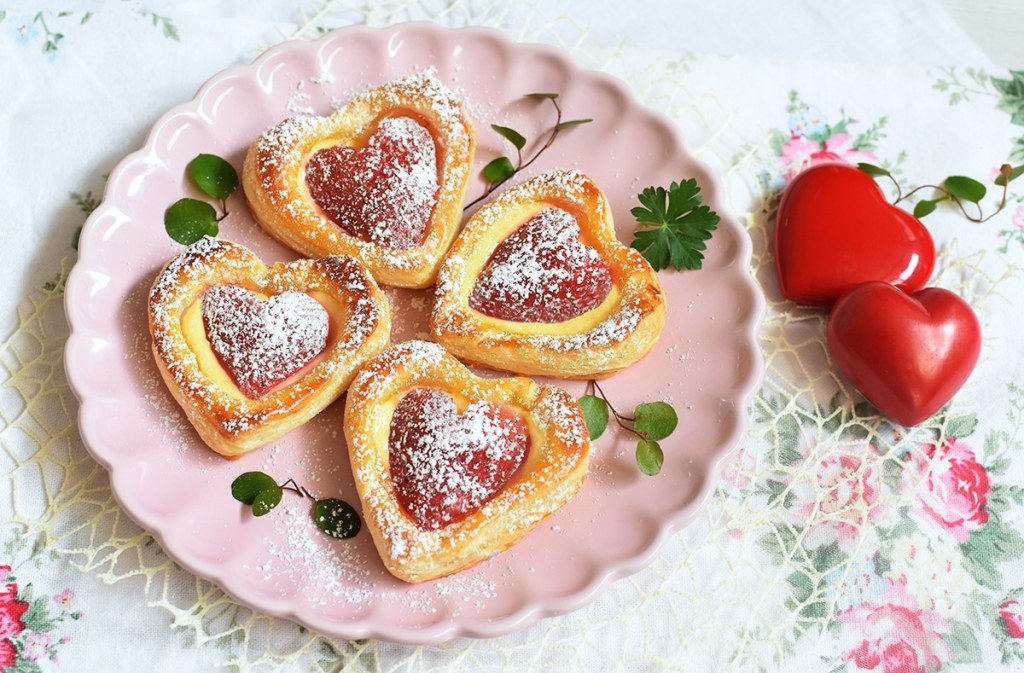 Vier Blätterteigherzen mit herzförmigen Erdbeeren als Füllung auf rosa Teller
