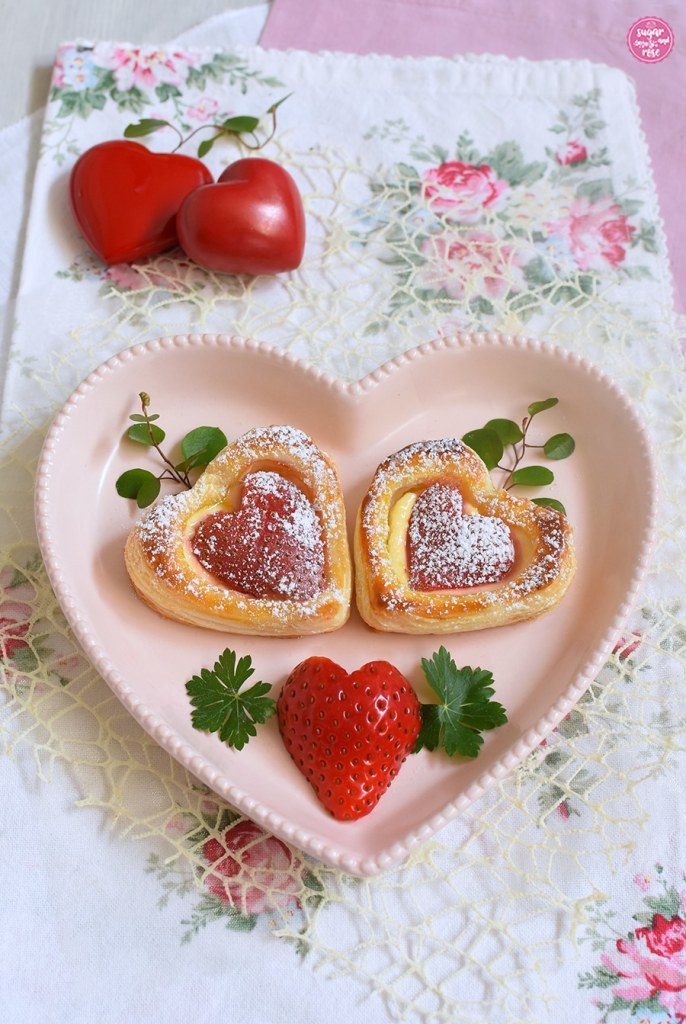 Zwei Erdbeerherzen auf herzförmigen lachsrosa Teller, dekoriert mit einer herzförmigen Erdbeere und zwei roten Dekoherzen