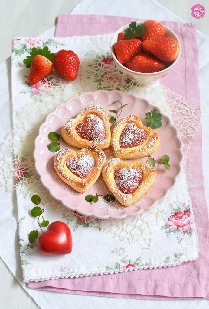 Vier Stück Erdbeerherzen auf rundem, rosa Keramik-Dessertteller, dekoriert mit kleinen grünen Blättern, dahinter ein kleines Schälchen mit Erdbeeren, alles auf Stoffservietten, einmal rosa, einmal mit Rosendekor