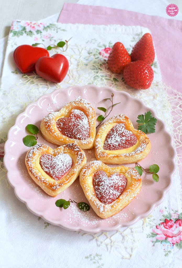 Vier Erdbeerherzen auf rosa Keramikteller angerichtet, im Hintergrund zwei rote Steinherzen und drei Erdbeeren