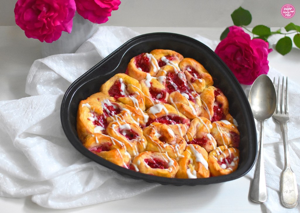 Himbeer-Plunder schneckenförmig aufgerollt und in einer Herz-Backform gebacken, anschließend mit etwas Zuckerguss übergossen. Im Vordergrund eine antike Gabel und ein ebensolcher Löffel sowie pinke Rosenblüten.