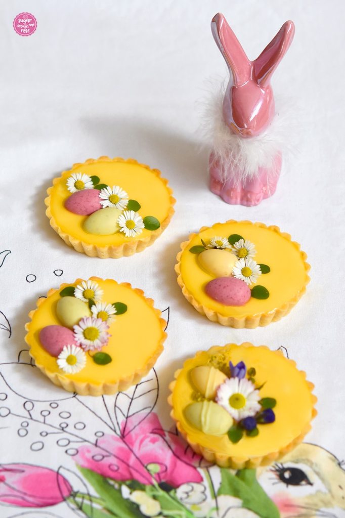 Vier Eierlikör-Tartelettes dekoriert mit Gänseblümchen, grünen Blättern und Veilchenblpten sowie bunten Schokoladeneiern. Im Hintergrund ein rosa Porzellanhase mit weißer Fell-Halskrause