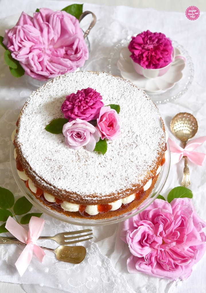 Victoria Sponge Cake auf Vintage-Glasteller, dekoriert mit drei unterschiedlich rosafarbenen Rosenblüten und zwei großen Blütn der Rose Liparfüm