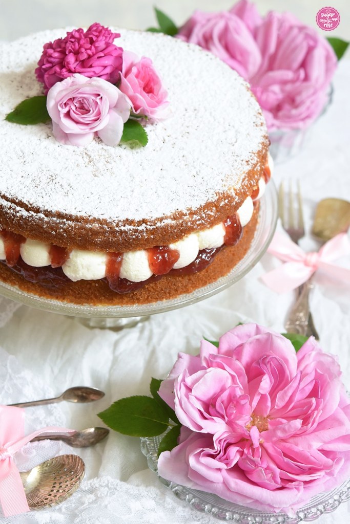 Victoria Spong Cake auf Glasteller mit Fuß, die leuchtend rote Erdbeermarmelade quillen zwischen den dicken Sahnetupfen heraus.