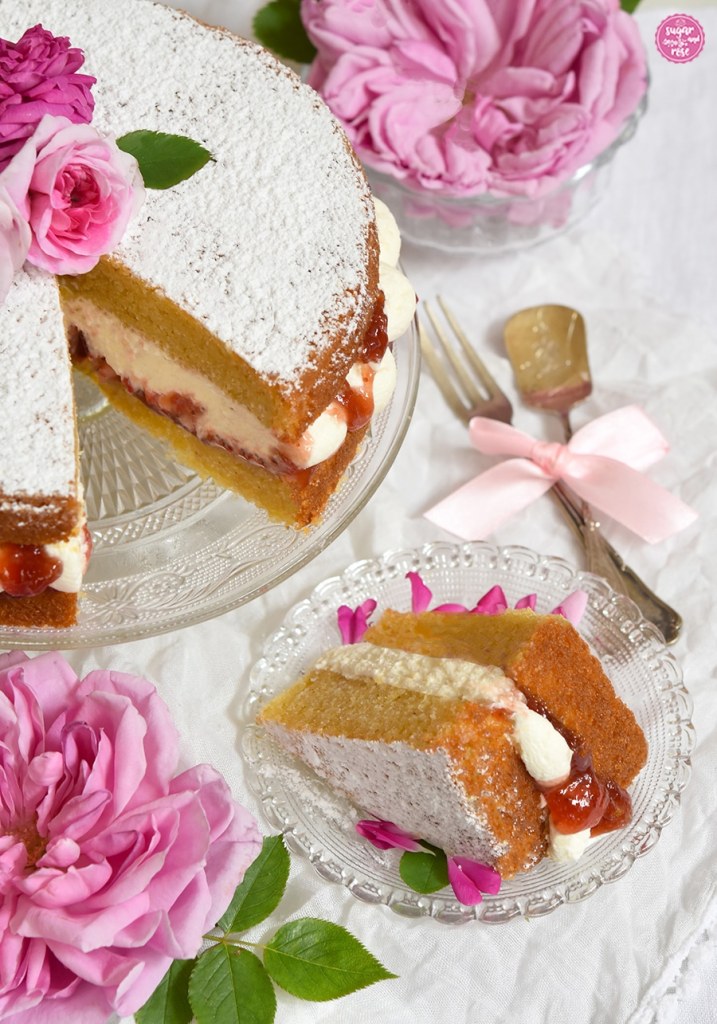 Großaufnahe eines Kuchenstücks vom Victoria Sandwich mit dicker Sahneschicht und Erdbeermarmelade, hinten die angeschnittene Torte und rosa Rosenblüten in kleinen Glasschalen