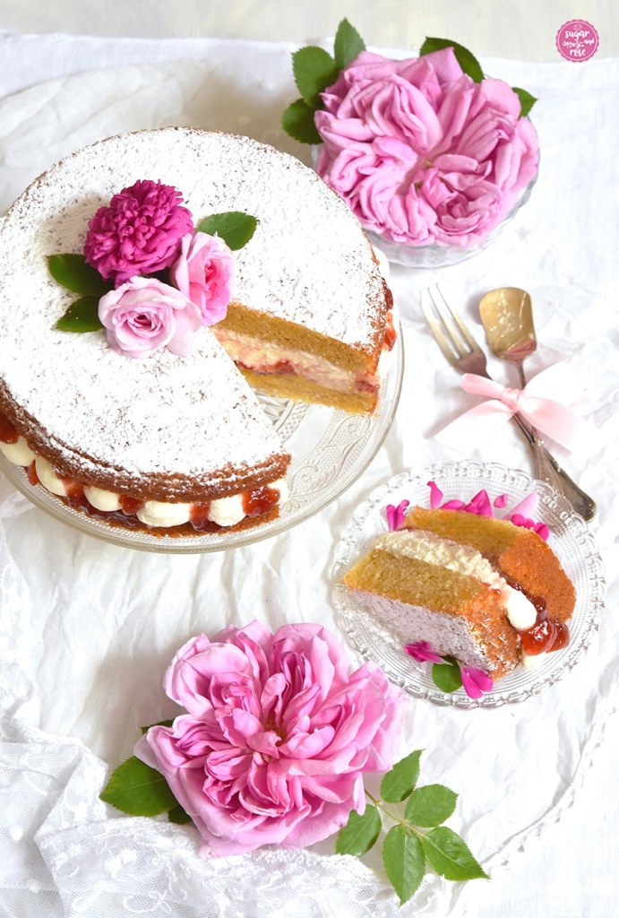 Angeschnittener Victoria Sponge Cake, ein Stück liegt auf einem kleinen Glastellerchen, rundum pinke Rosenblütenblätter, daneben zwei rosa Rosenblüten und Silberlöffel und Silbergabel mit rosa Schleife