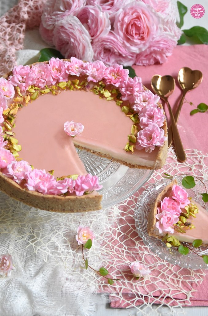 Angeschnittene Rosen-Panna-cotta-Tarte auf Vintage-Glasteller, ein Stück auf kleinem Glasteller davor, im Hintergrund zwei herzförmige rose-gold-farbene Löffel