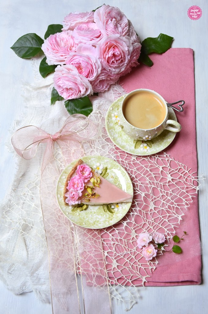 Ein Stück der Rosen-Panna-cotta-Tarte auf einem hellgrünen Dessertteller mit weiß-grünen Blüten, dahinter eine dazu passende Kaffeetasse mit Kaffee, beides auf einer rosa Leinenserviette und einem transparenten cremfarbenen Deckchen, eine transparente breite rosa Schleife und ein ein Strauß rosa Rosen