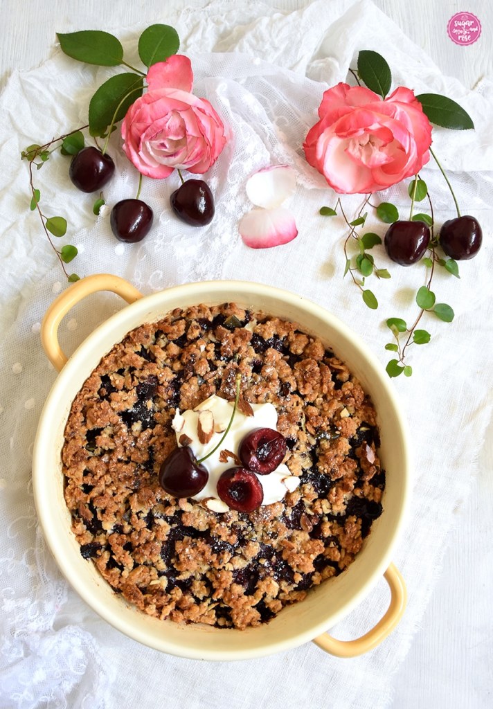 Kirsch-Crumble in runder gelber Emaillepfanne, dahinter zwei Rosenblüten und Kirschen
