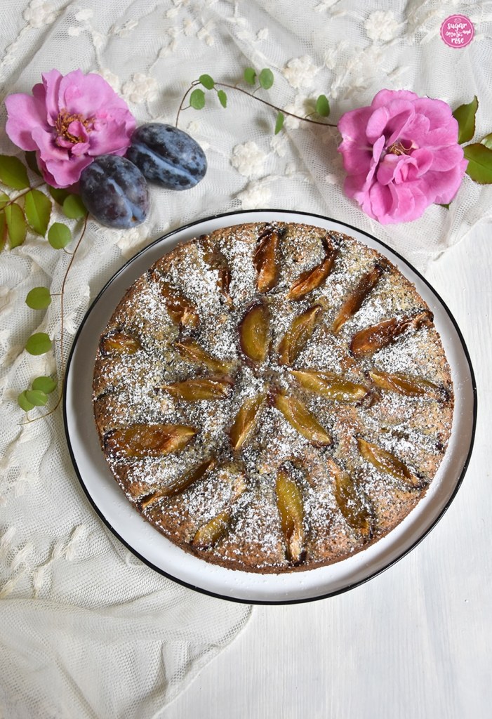 Runder Mohnkuchen mit aufgefächerten Zwetschken obenauf, bestreut mit Puderzucker, im Hintergrund zwei blaue Zwetschken und zwei rosa Rosenblüten