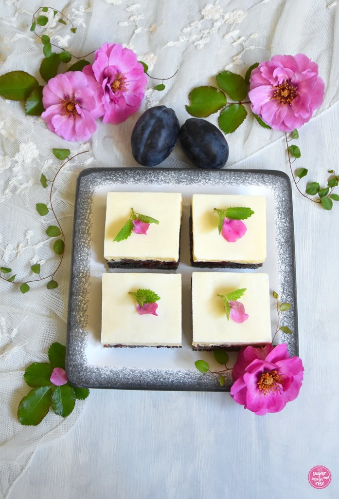 Mohn-Zwetschken-Kuchen mit Vanillecreme auf eckigem weißen Gmundner Teller mit grauem Rand, im Hintergrund rosa Blüten der Rosa Orienta Djamila und zwei Zwetschken