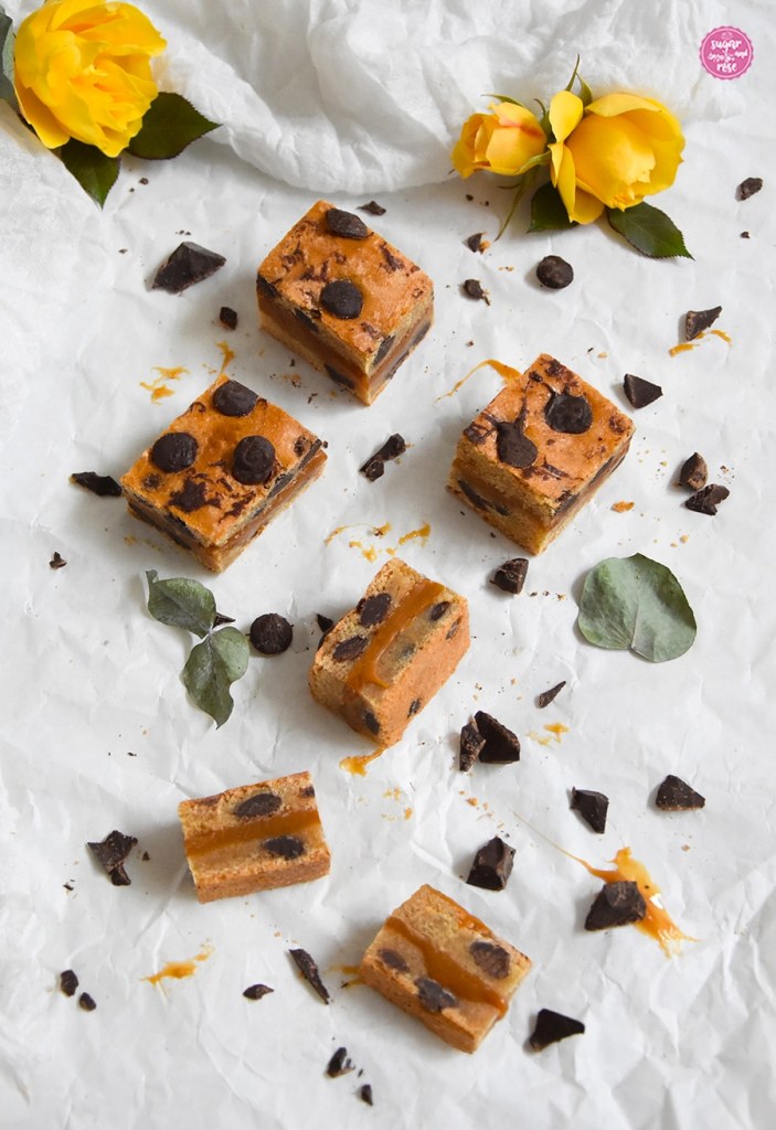 Karamell-Cookies auf Butterpapier, teils in große Riegel geschnitten, teils in kleine Stücke, dekoriert mit zwei Blüten der gelben Rose Tequila Gold