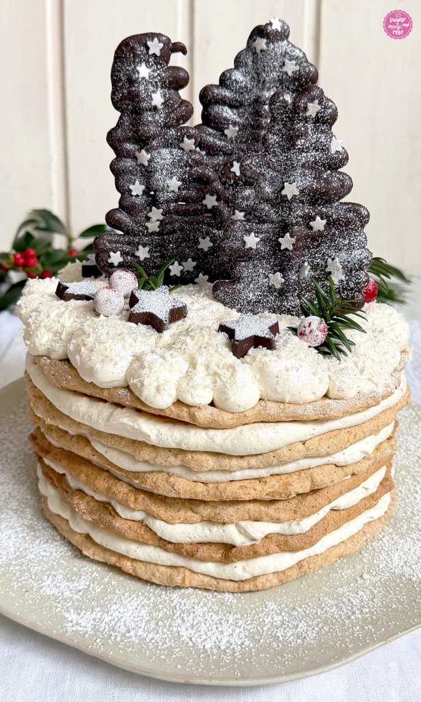Wintertorte mit sechs hellbraunen Nussbaiserböden und dicker weißer Creme, dekoriert mit drei dunklen Schokoladebäumchen und Schokosternen mit Silberglitzer