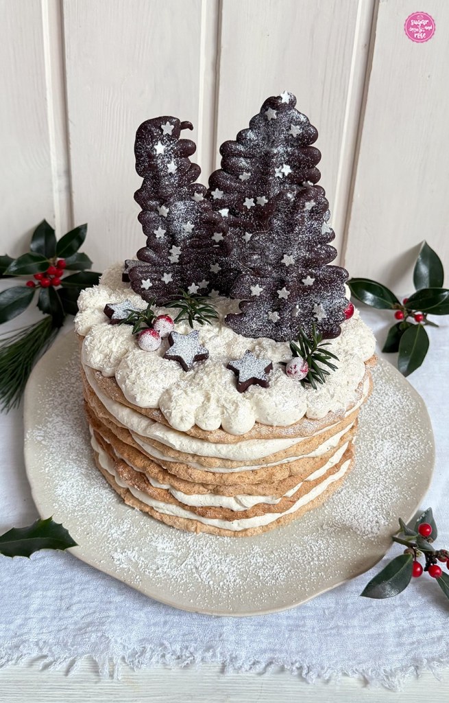 Wintertorte mit Dulce de Leche und Schokobäumchen und Schokosternen vor weißer Holzwand, dekoriert mit Zweigen von grüner Stechpalme mit roten Früchten